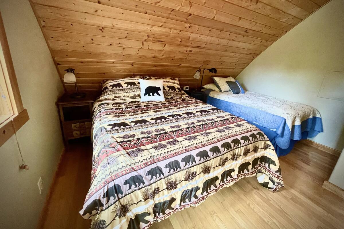 Un dormitorio con techo inclinado, una cama grande con manta estampada, dos mesitas de noche y una mesita auxiliar.