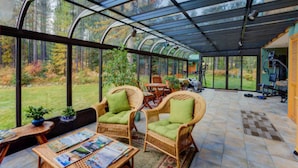 Une véranda meublée de mobilier en osier, dotée d’une verrière et offrant une vue sur une forêt.
