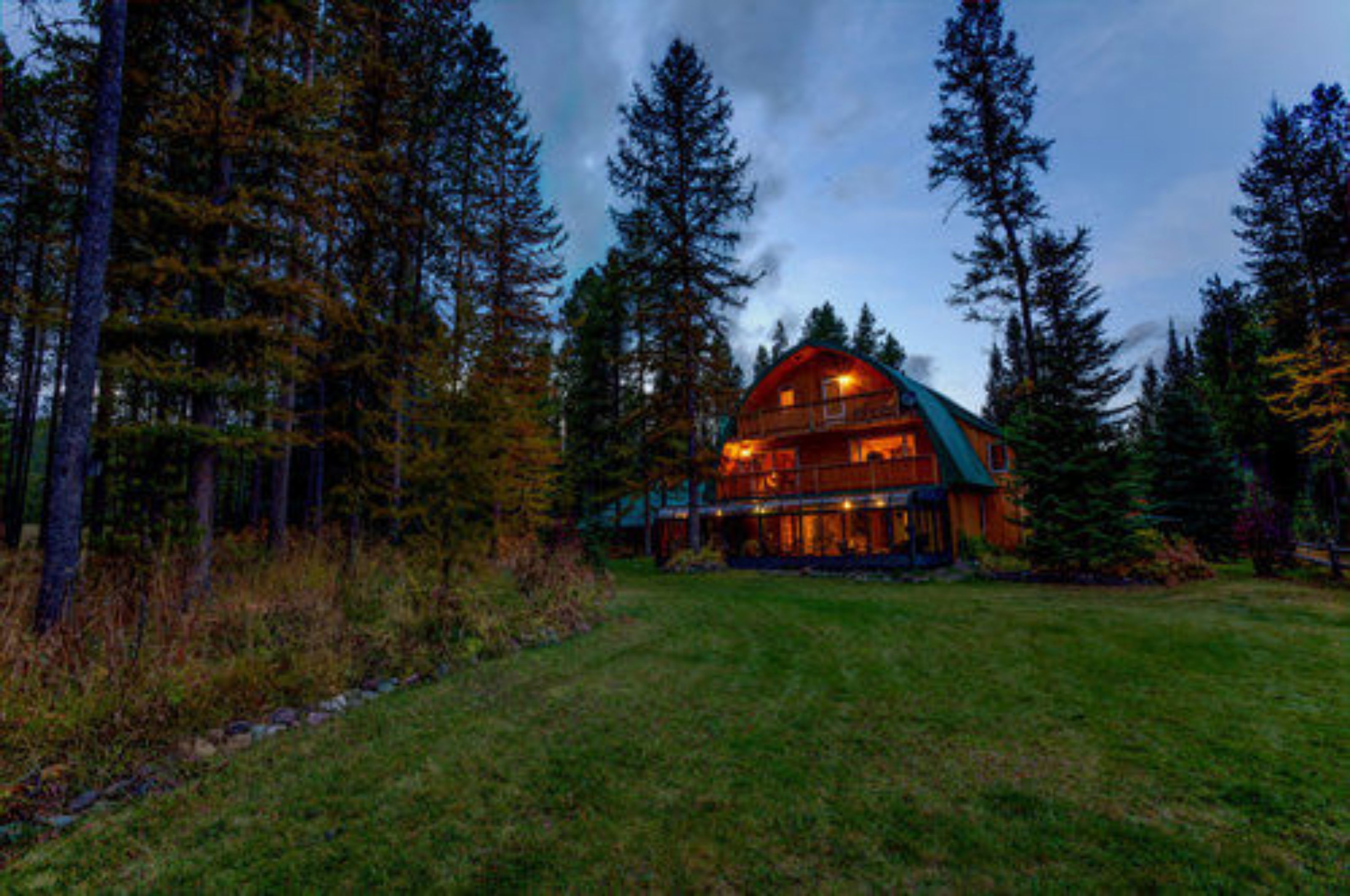 A log cabin with a large porch and multiple windows, surrounded by tall pine trees.