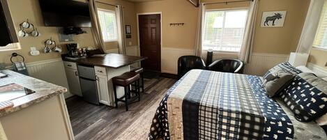 A compact, well-lit room with a kitchenette, a bed with a checkered blanket, and a mounted TV.