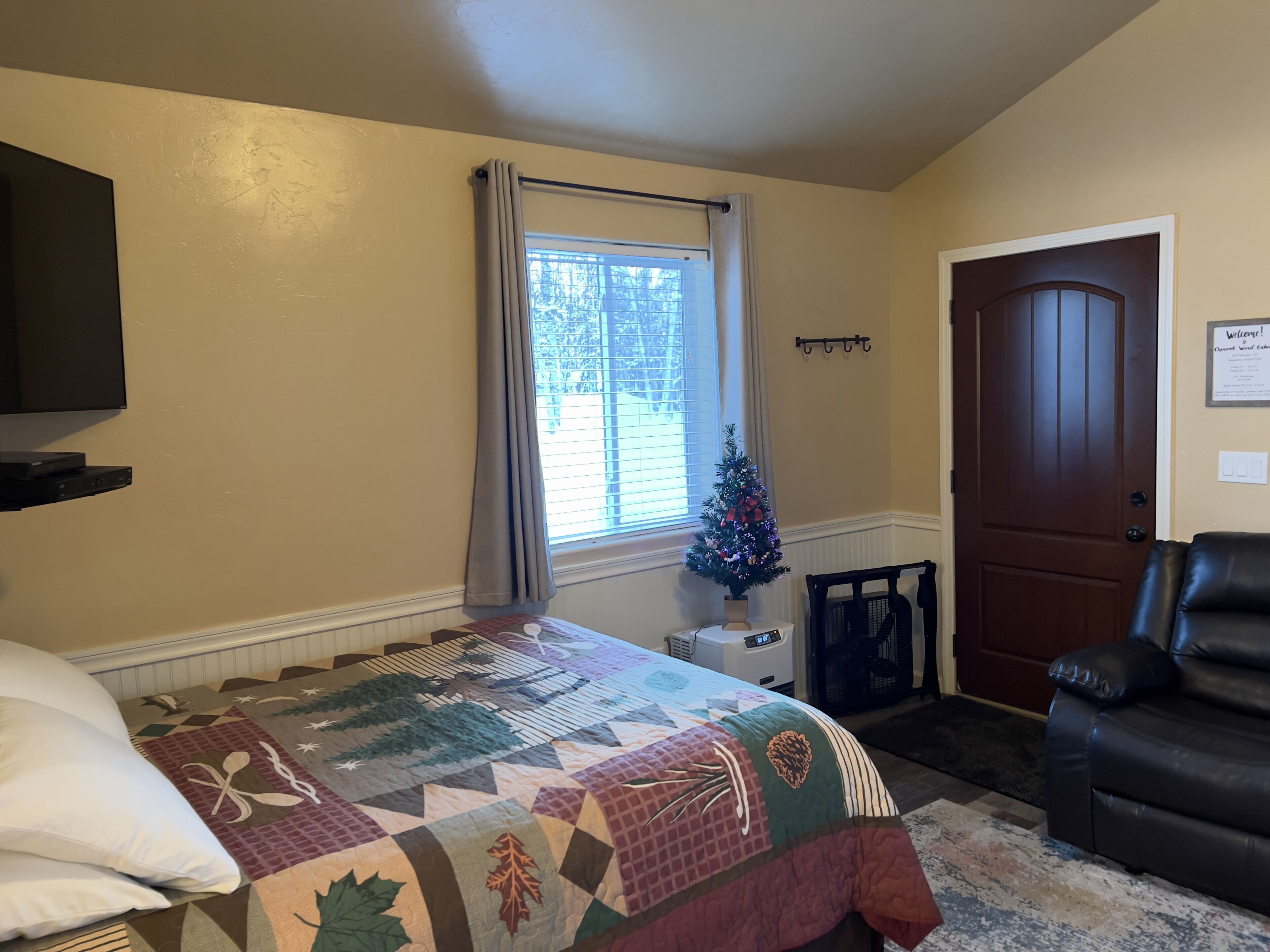 Ein Schlafzimmer mit einem Bett, einem Fernseher, einer kleinen Weihnachtstanne, einer Tür und einem Sessel.