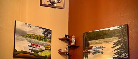 Quarto com uma prateleira de madeira que contém uma cesta, uma caneca e um pequeno dispositivo eletrônico. Há duas pinturas na parede, uma retratando uma paisagem com um avião vermelho e outra mostrando uma cena de floresta com um barco. Uma luminária suspensa com três lâmpadas é visível acima da prateleira.