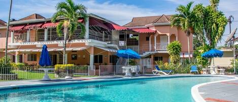 A swimming pool with lounge chairs and umbrellas, surrounded by buildings with balconies and palm trees.