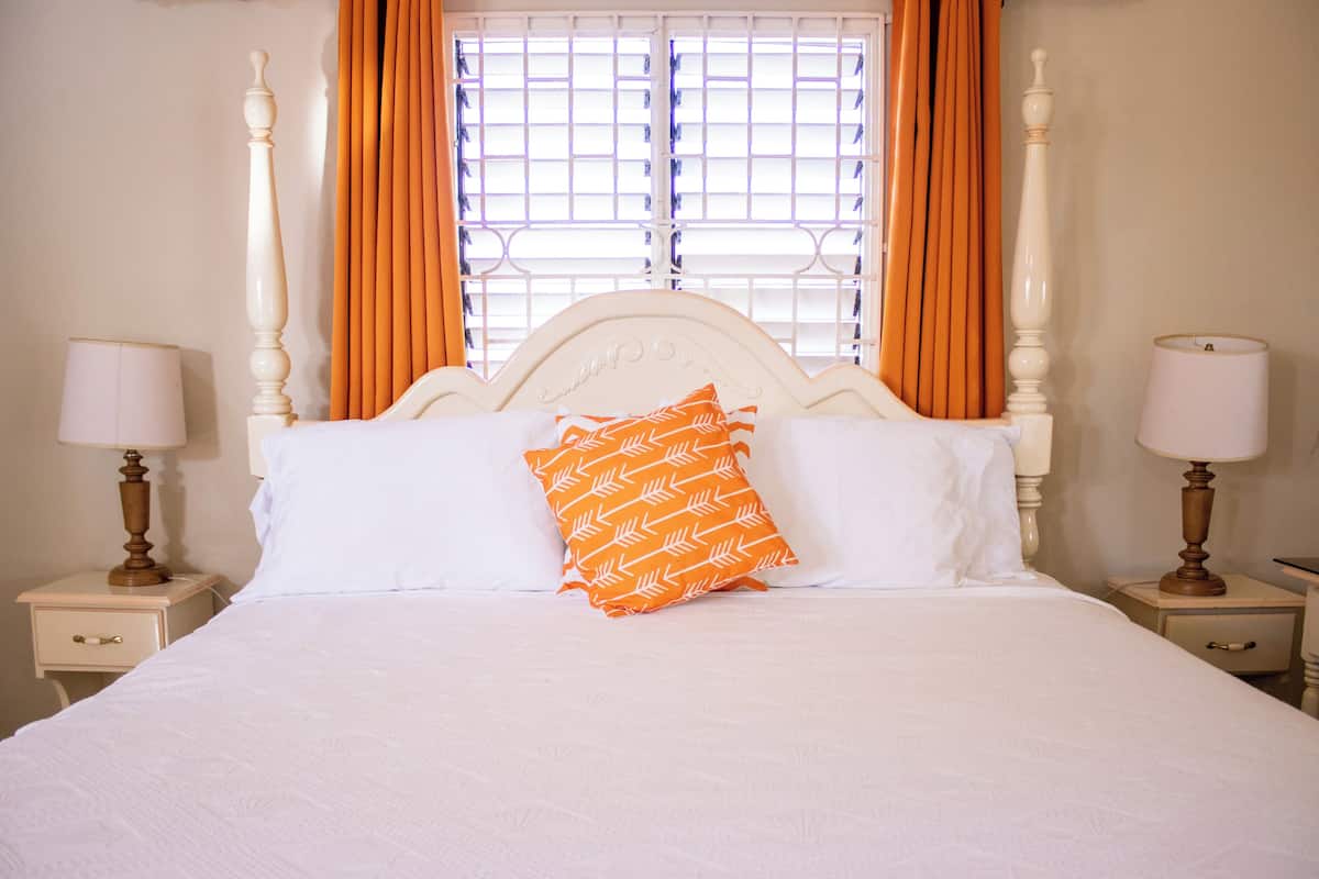 Un lit bien fait, avec une literie blanche et un coussin à motifs orange, flanqué de deux tables de chevet avec des lampes, dans une pièce dotée d’une fenêtre et de rideaux.