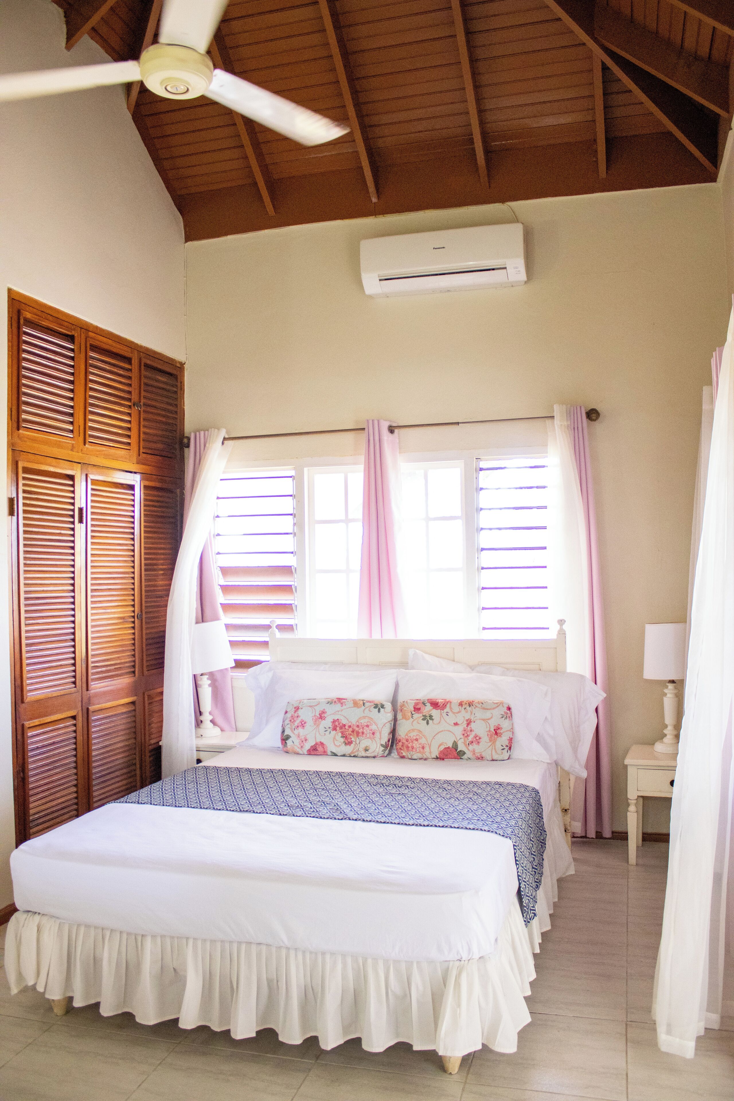 Un dormitorio con cama, ventilador de techo, unidad de aire acondicionado, contraventanas de madera y una ventana con cortinas.