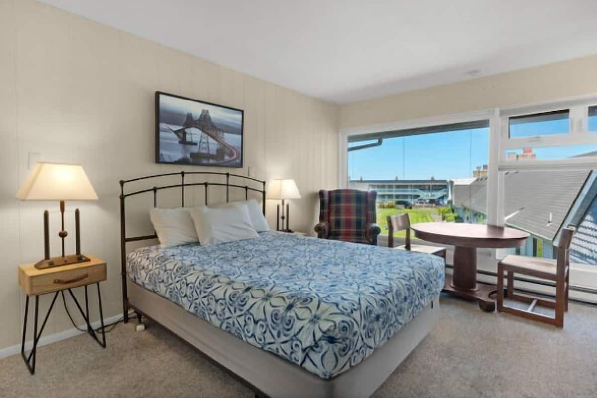 A hotel room with a bed, two bedside lamps, a chair, a small table, and a balcony with a view of a bridge.