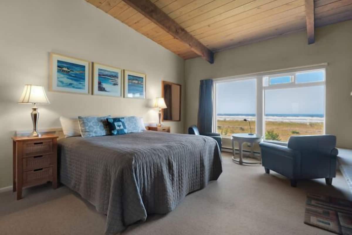 A bedroom with a large bed, a small table, a chair, and a view of the beach.