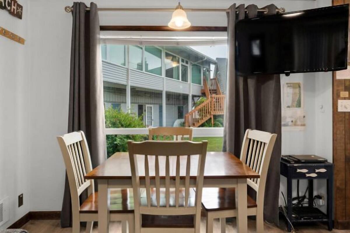 A dining area with a wooden table and chairs, a television on the wall, and a view of a building outside.