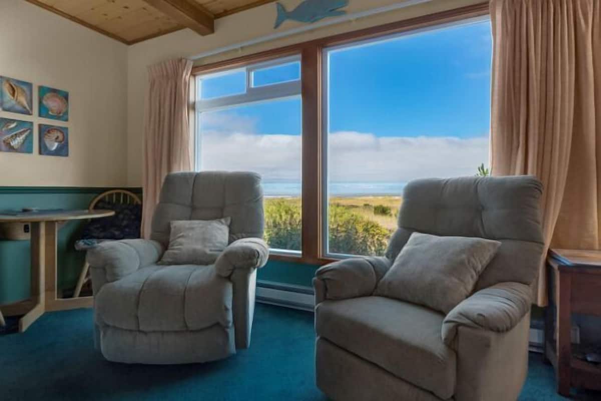A room with two beige armchairs facing a large window with a view of the beach and sky.