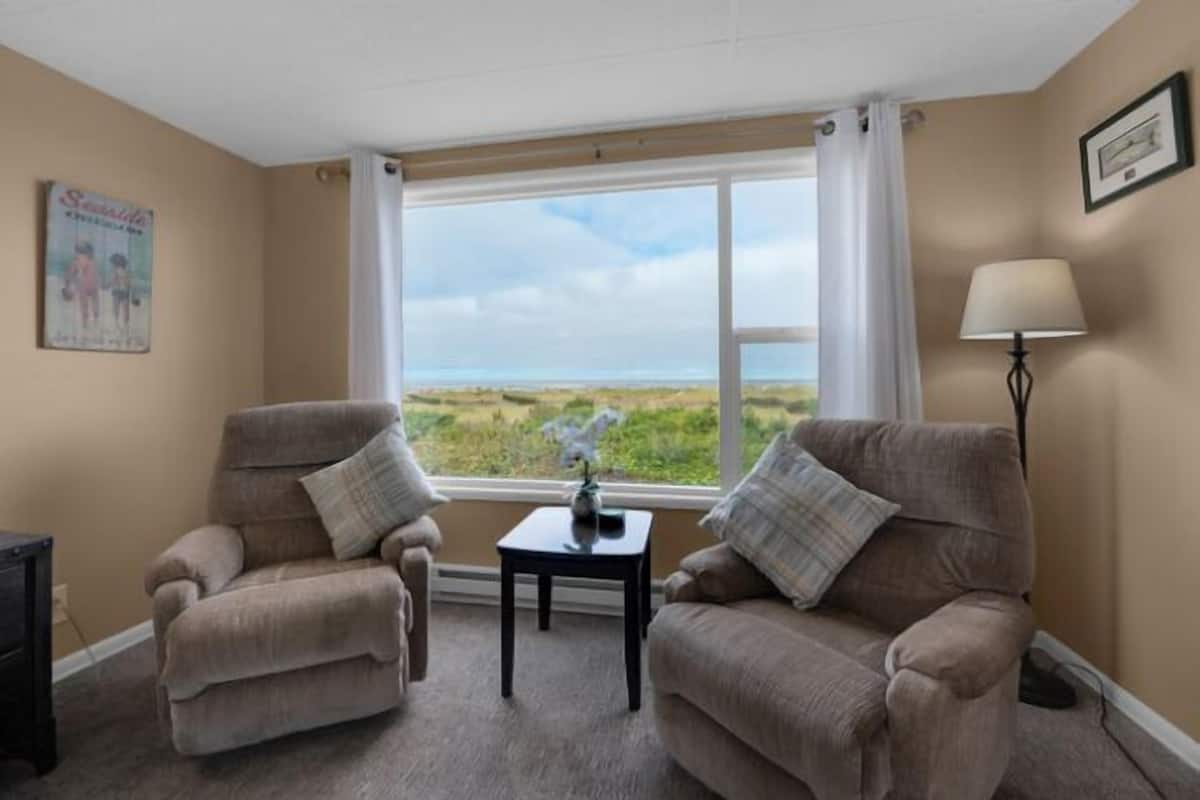 A room with two beige armchairs, a small table with a vase, a lamp, and a window with a view of the beach.