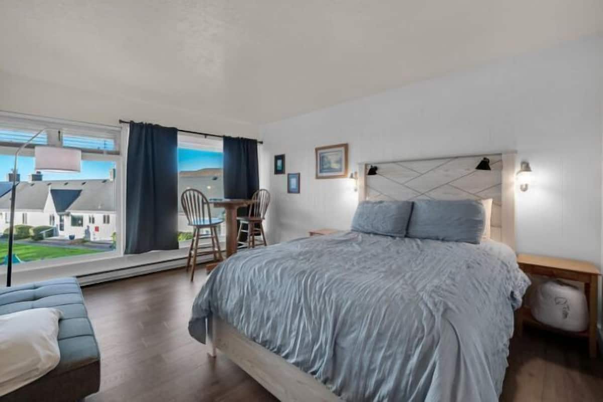 A bedroom with a large bed, a desk, and a chair. There is a window with curtains, a lamp, and a picture on the wall.