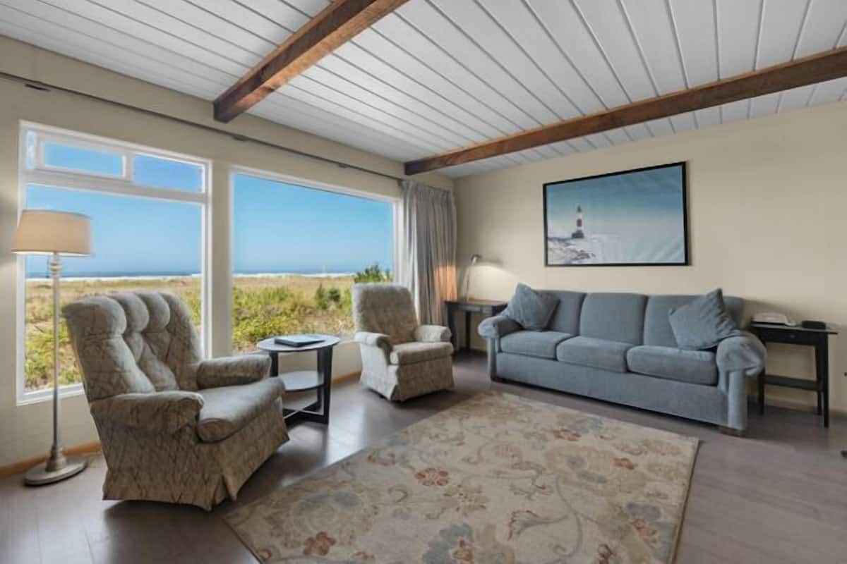A living room with a view of the beach, a sofa, two armchairs, a coffee table, and a lamp.