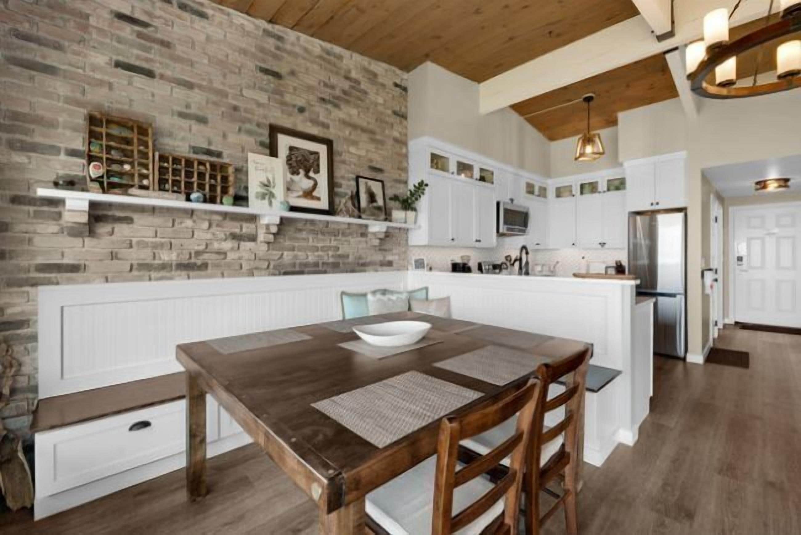 A kitchen with a brick wall, white cabinetry, and a wooden dining table with a bowl.