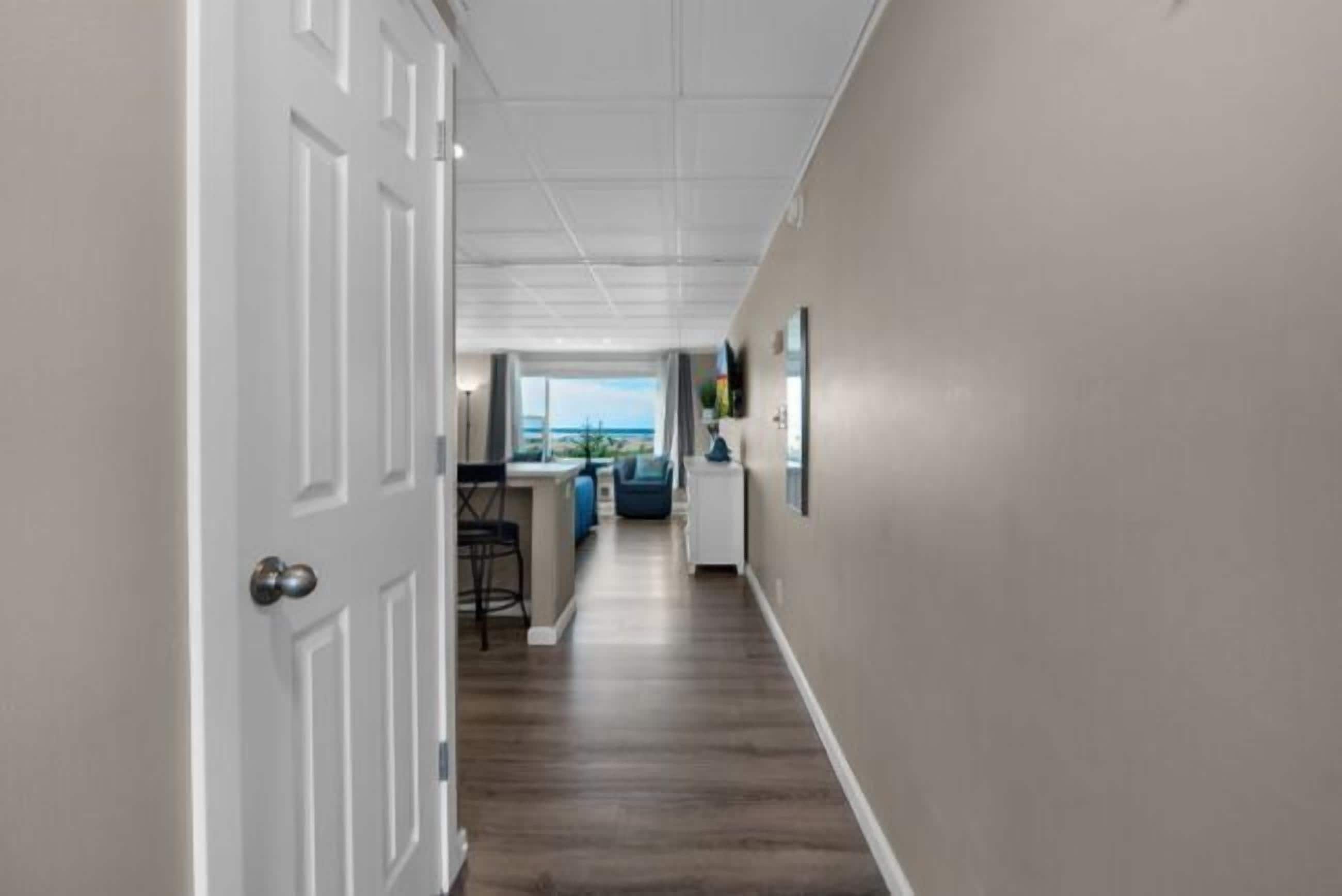 A hallway with a white door, a kitchen area with a view, and a dining area with a blue chair.