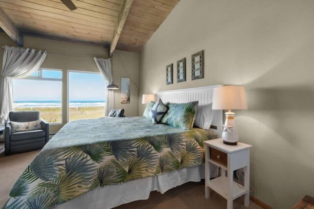 A bedroom with a large bed, a nightstand with a lamp, a chair, and a view of the beach through the window.