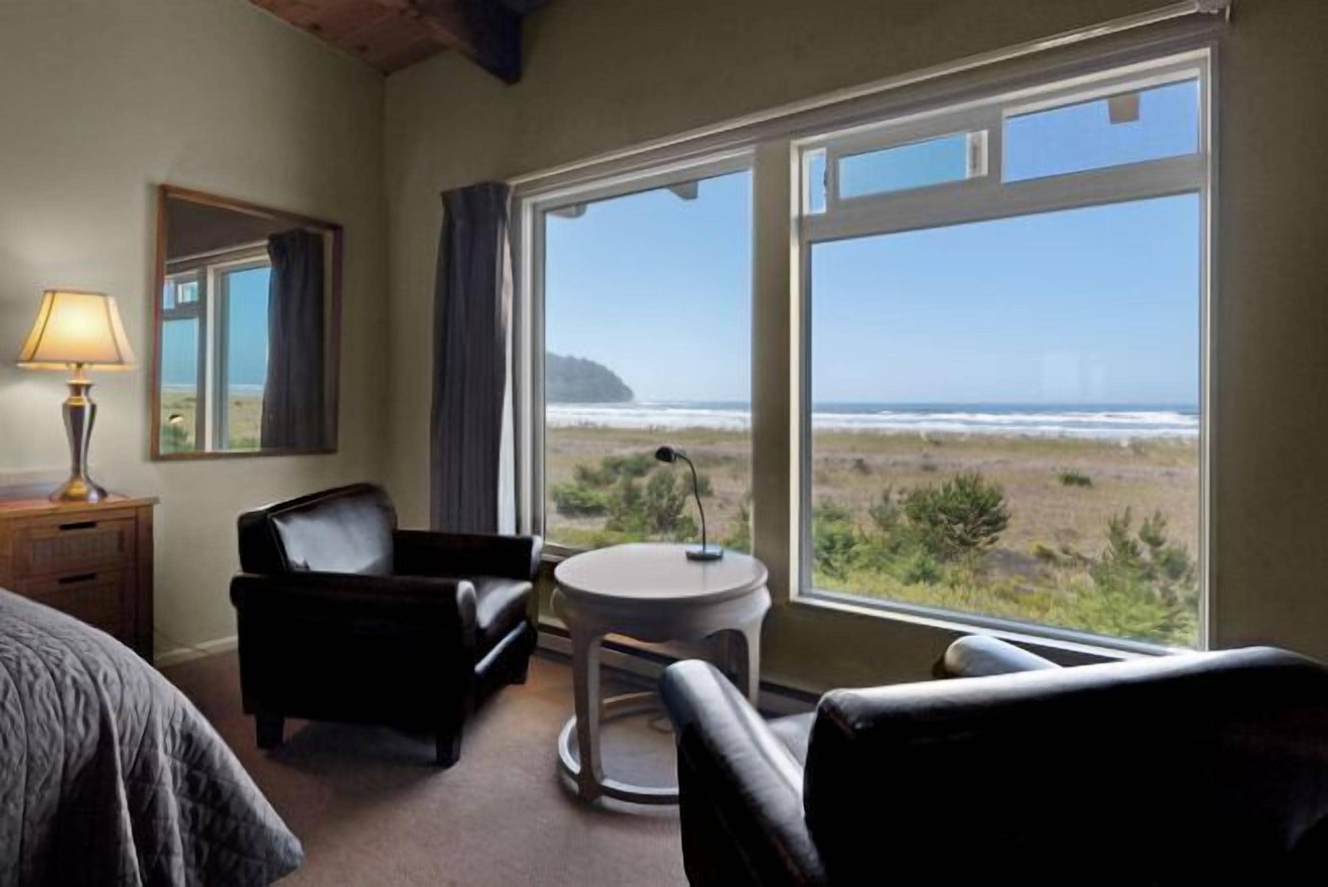 A room with a bed, a desk, a chair, a small table, and a large window offering a view of the beach and ocean.