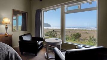 A room with a bed, a desk, a chair, a small table, and a large window offering a view of the beach and ocean.