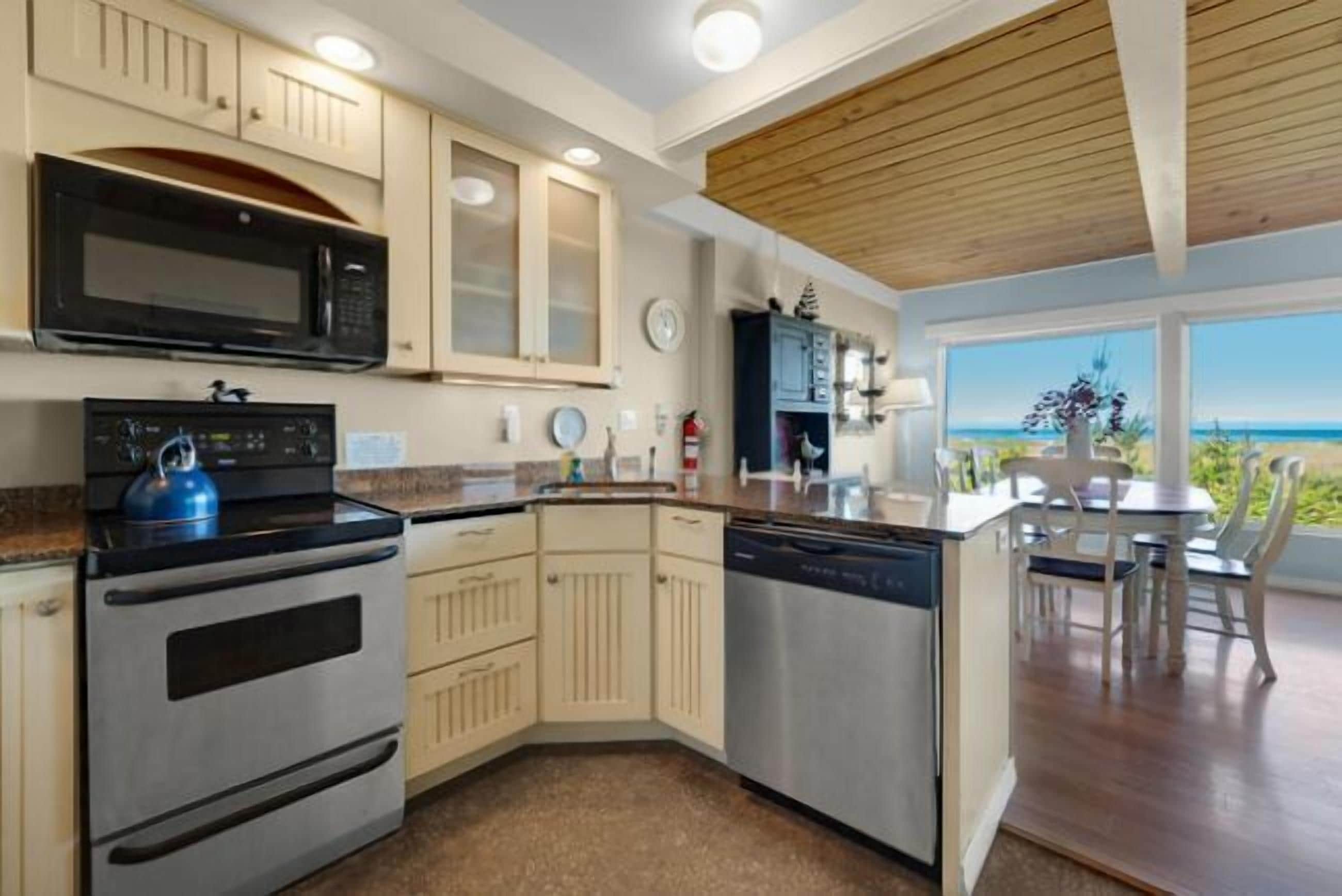 A modern kitchen with a built-in oven, microwave, and dishwasher. It features light-colored cabinetry, a wooden ceiling, and a view of the ocean through large windows.