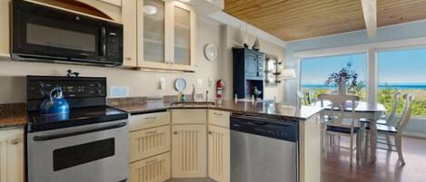 A modern kitchen with a built-in oven, microwave, and dishwasher. It features light-colored cabinetry, a wooden ceiling, and a view of the ocean through large windows.