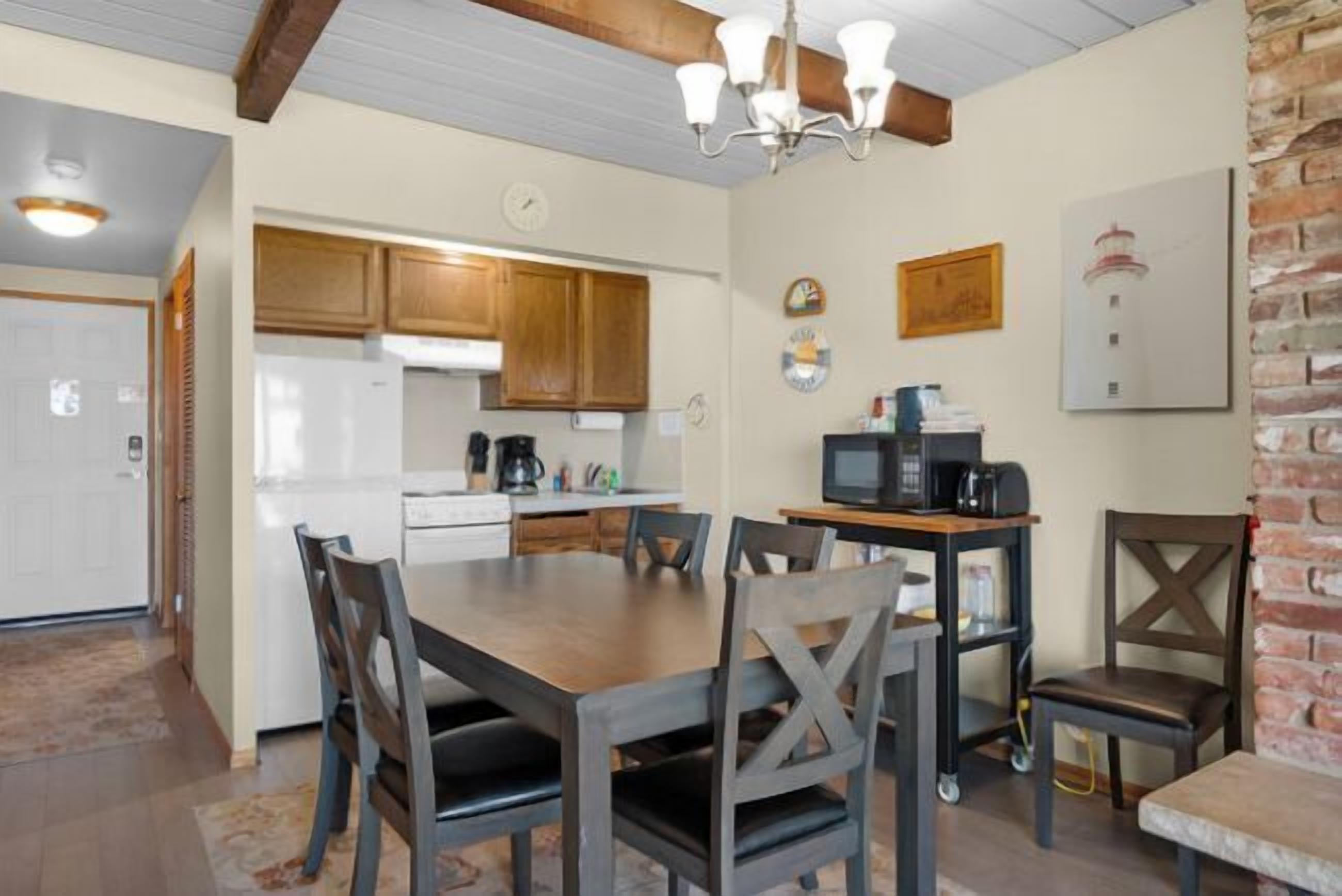 A dining area with a wooden table and chairs, a kitchen in the background, and a brick wall.