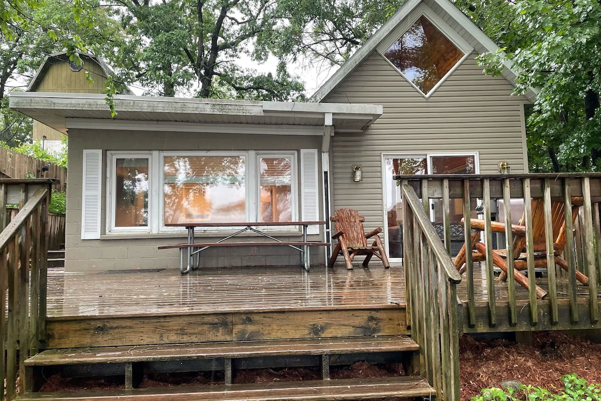 A wooden deck with a bench and a chair, a small house with a porch, and a wooden fence.