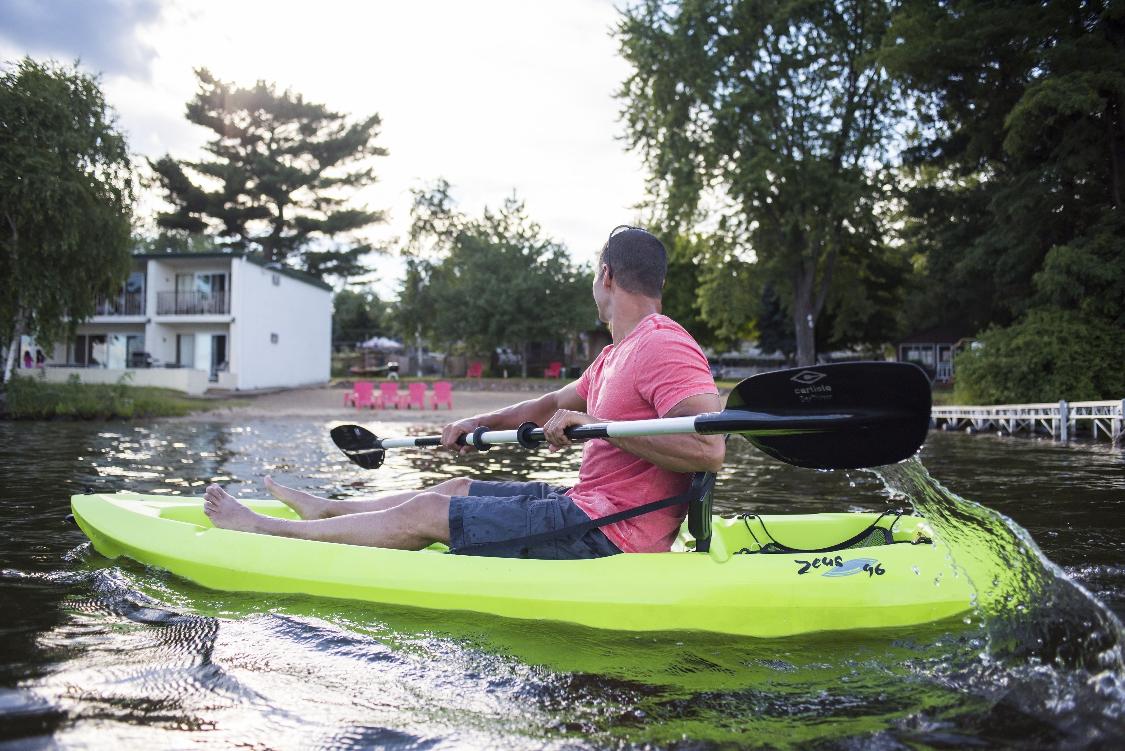 Una persona haciendo kayak en un kayak verde sobre un cuerpo de agua, con árboles y un edificio al fondo.