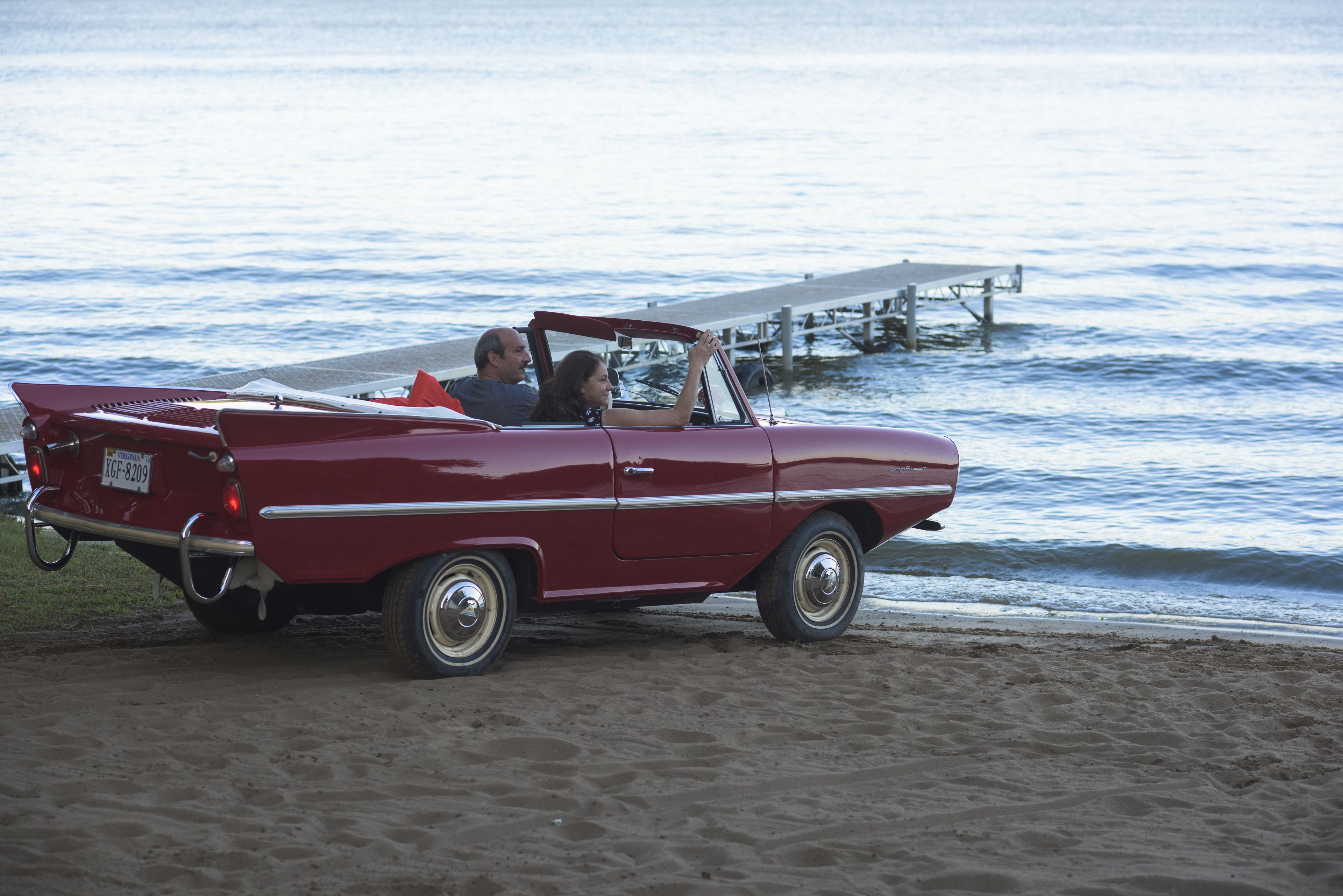 Une voiture rouge décapotable d’époque est garée sur une plage de sable, avec une jetée en arrière-plan.