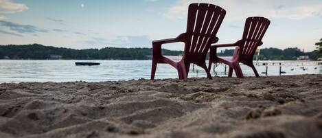 To Adirondack-stole på en sandstrand med en sø og skov i baggrunden.
