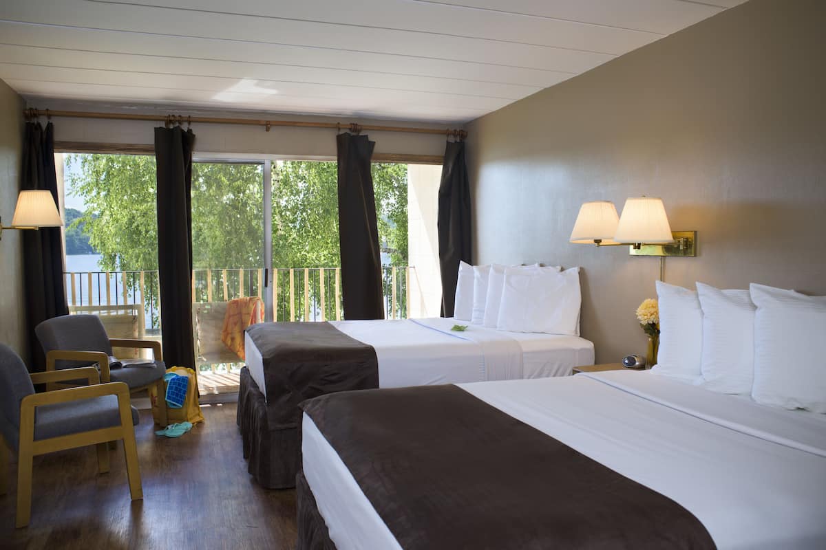 A hotel room with two beds, a chair, a small table, and a balcony with curtains.