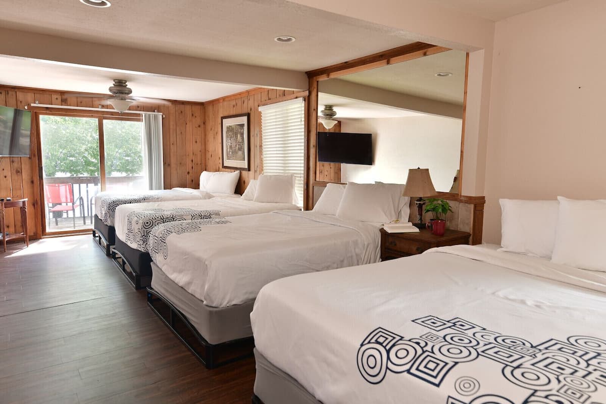 A hotel room with three beds, wooden flooring, a large mirror, a TV, and a balcony with a red chair.