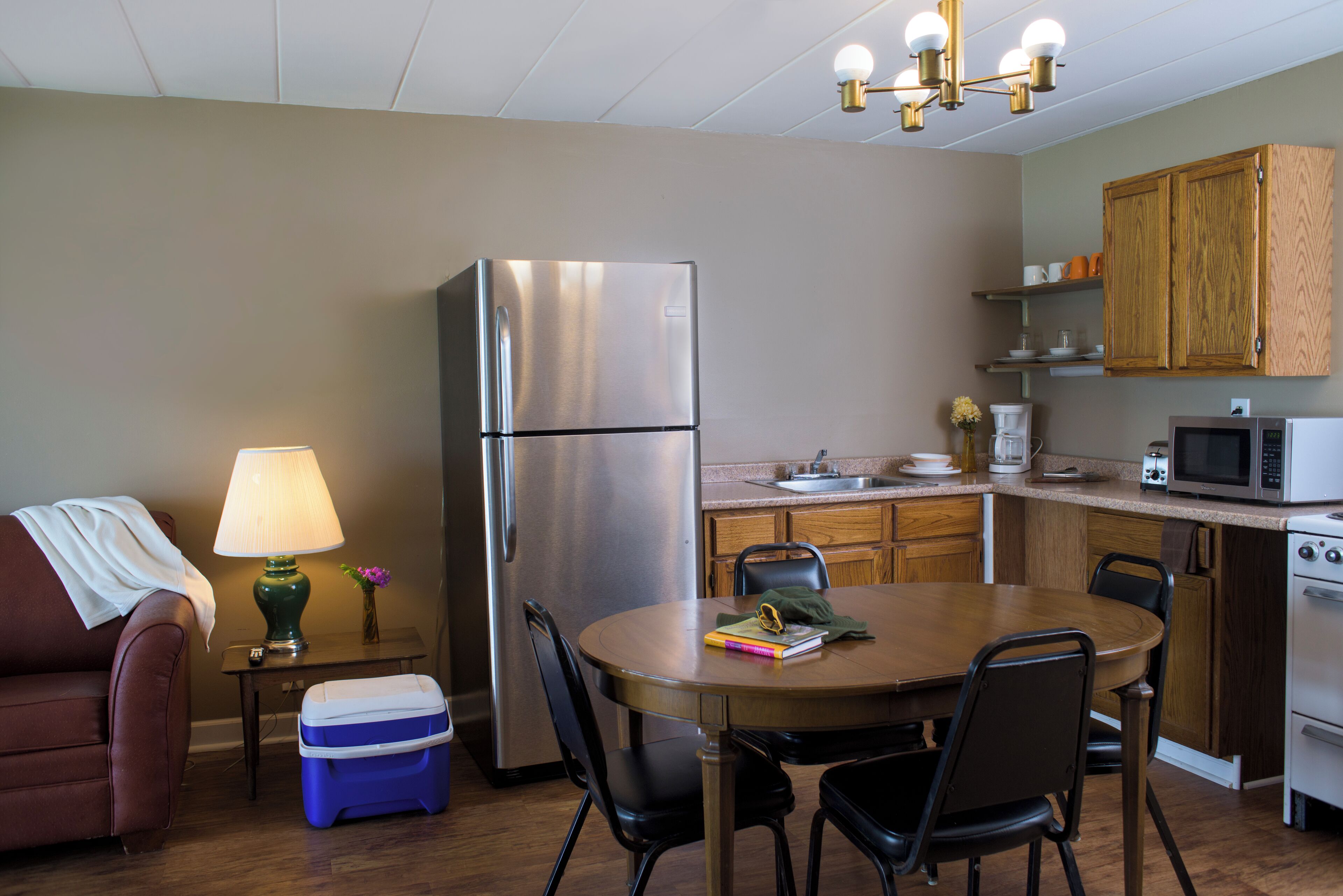A compact kitchen with a refrigerator, microwave, and stove. There is a dining table with chairs, a small side table with a lamp, and a couch.