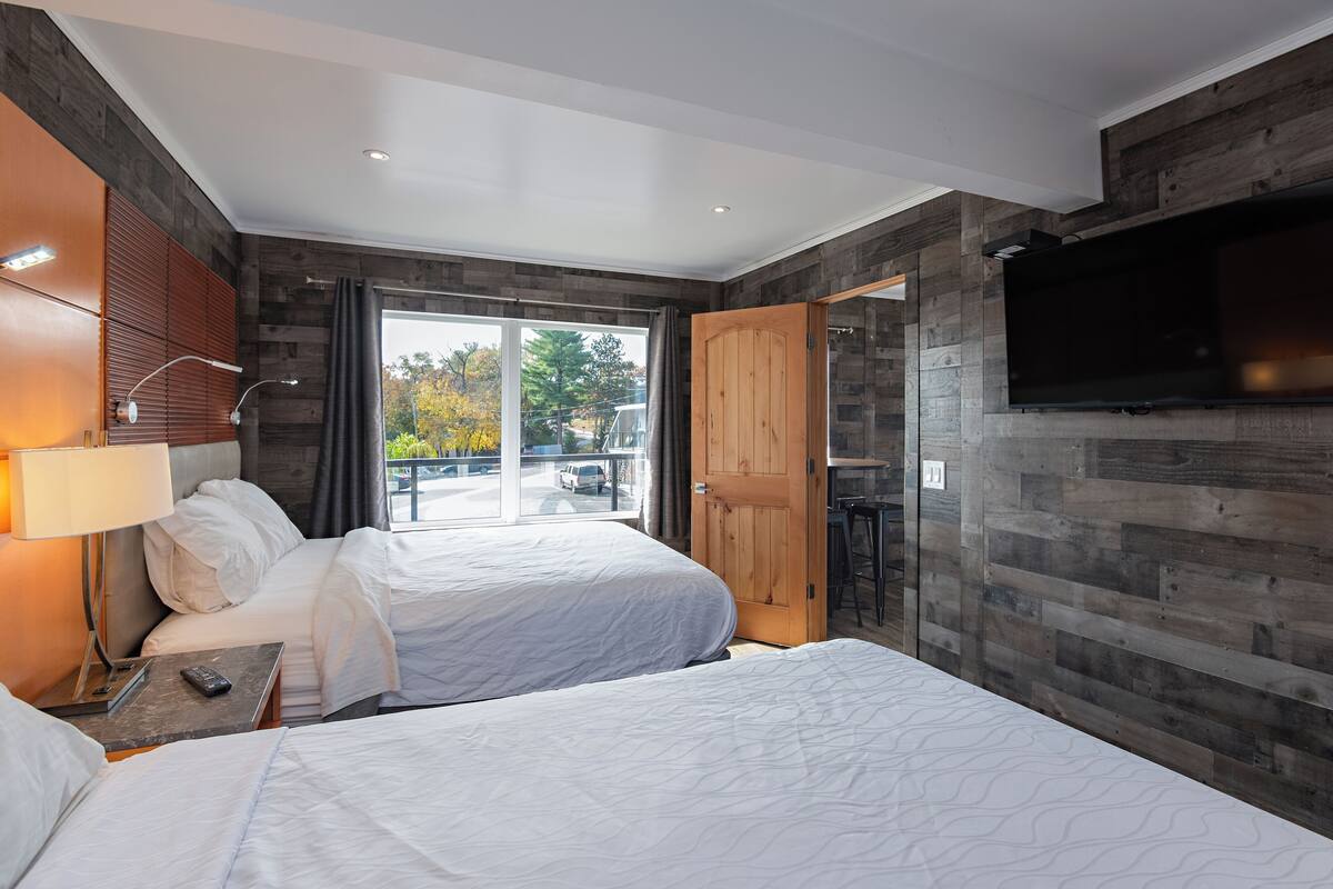 A hotel room with two beds, a TV, a desk, and a large window with a view of the outside.