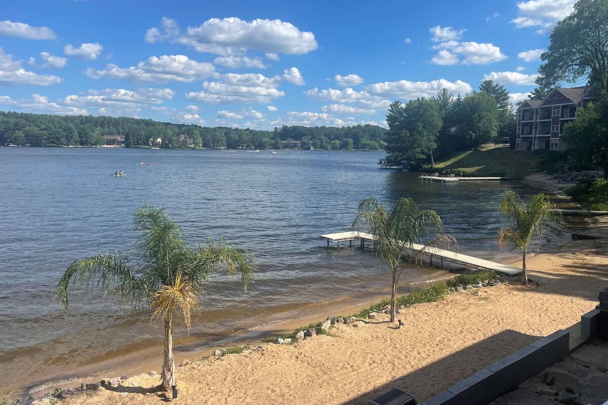 A lakeside view with a sandy beach, a dock, and a row of trees.