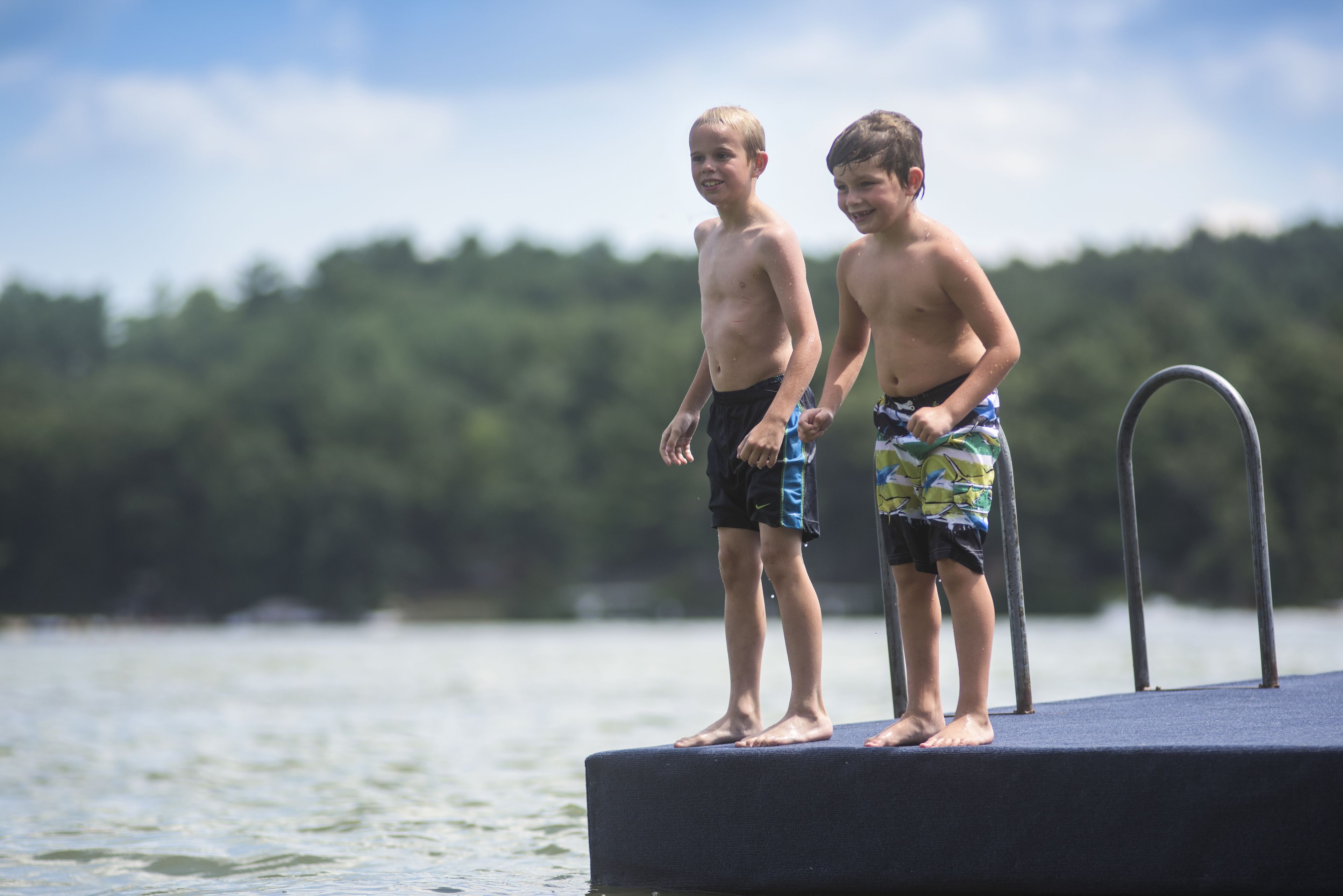 Two children standing on a dock by a lake with a forest in the background.