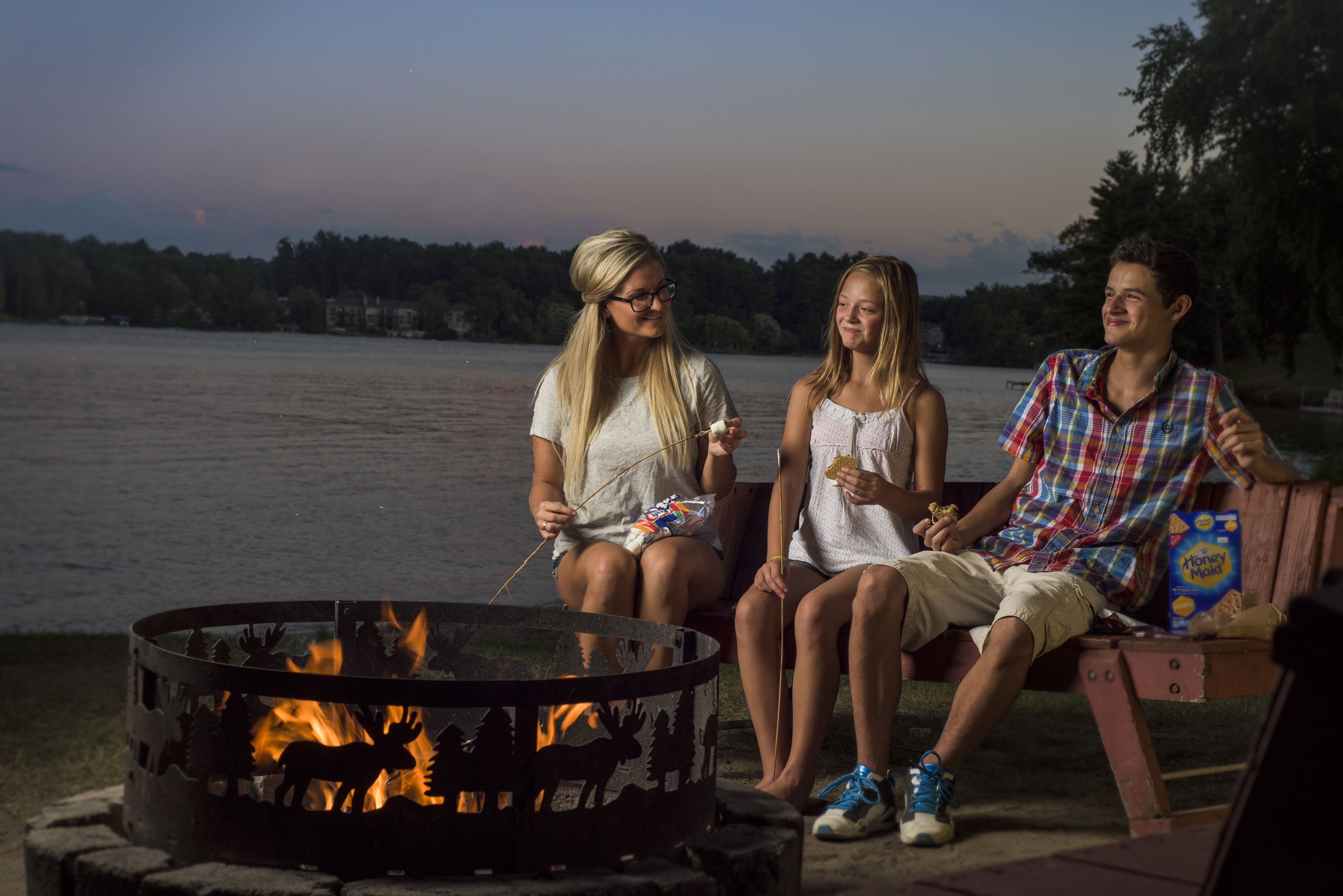 Three people sitting by a campfire with a lake and trees in the background.