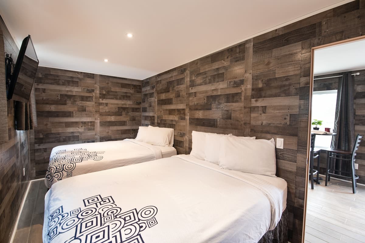 A hotel room with two beds, wooden panel walls, a flat-screen TV, and a dining area visible through an open door.