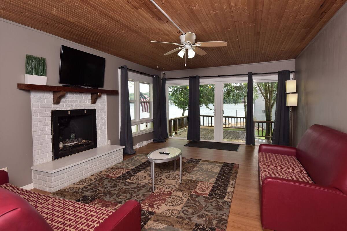 A spacious living room with a fireplace, red leather sofas, a ceiling fan, and a large window overlooking a deck.