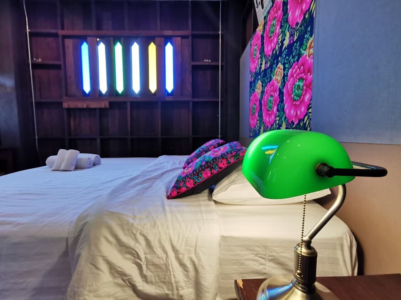 A hotel room with a bed, a green desk lamp, a wall with a floral pattern, and a window with vertical light fixtures.