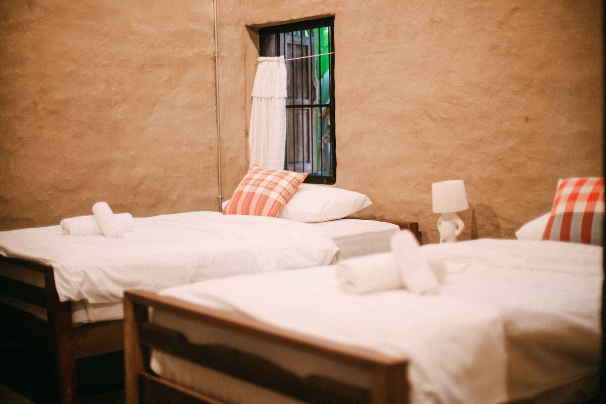A room with two single beds, each with a pillow and a rolled towel. There is a window with a curtain and a small table with a lamp.
