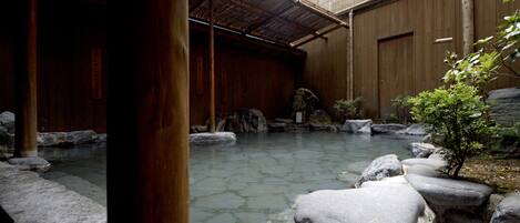 A traditional Japanese onsen with stone baths, wooden structures, and a small garden area with rocks and plants.