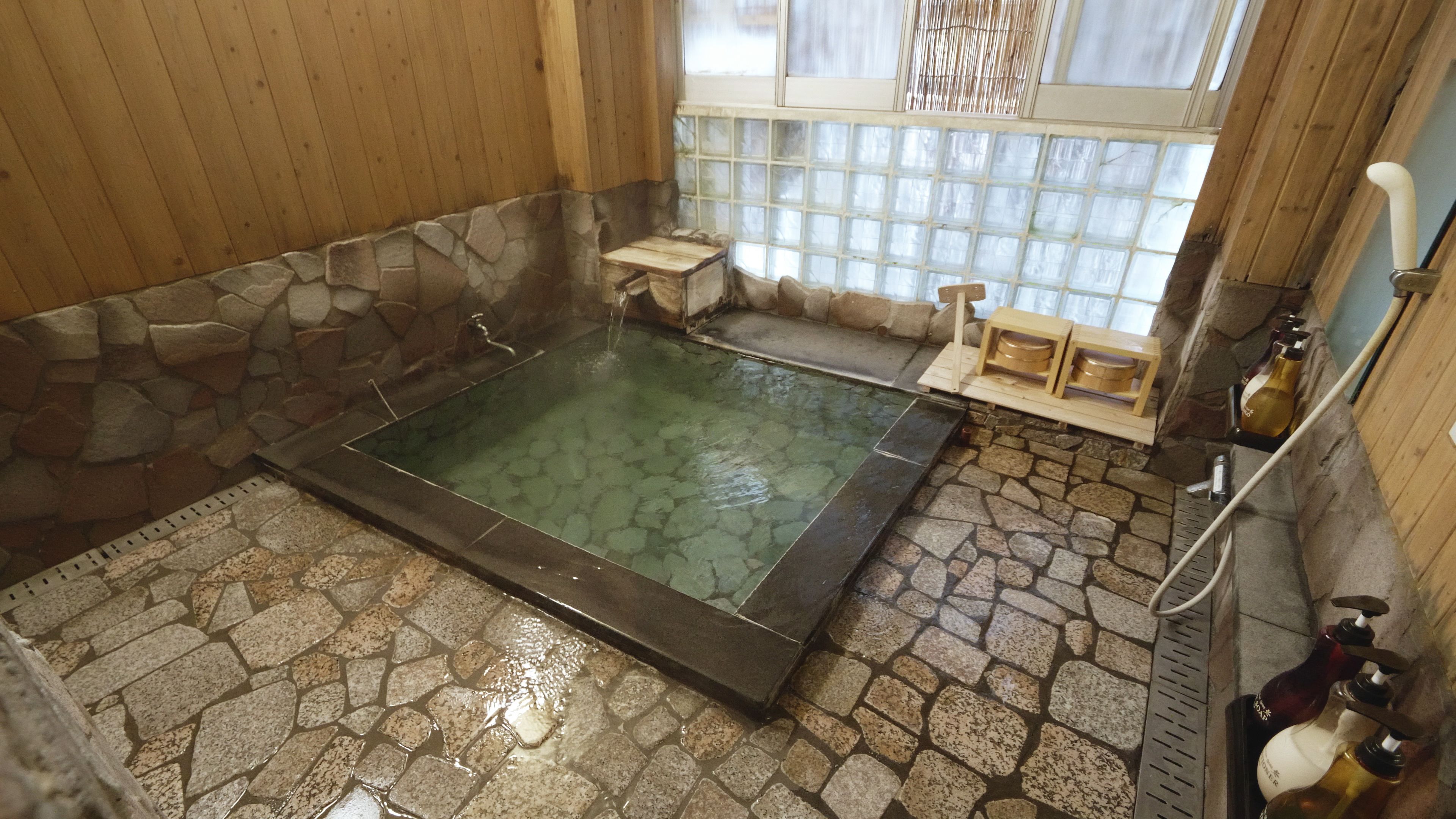 A stone-floored hot tub with a wooden bench and a small table, a stone wall, and a window with a view.