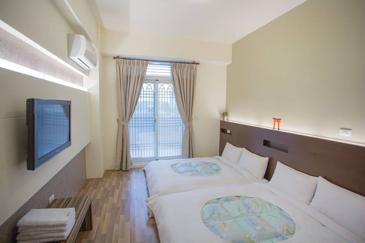 Hotelkamer met twee bedden, een aan de muur bevestigde televisie, een raam met gordijnen en een klein tafeltje met handdoeken.