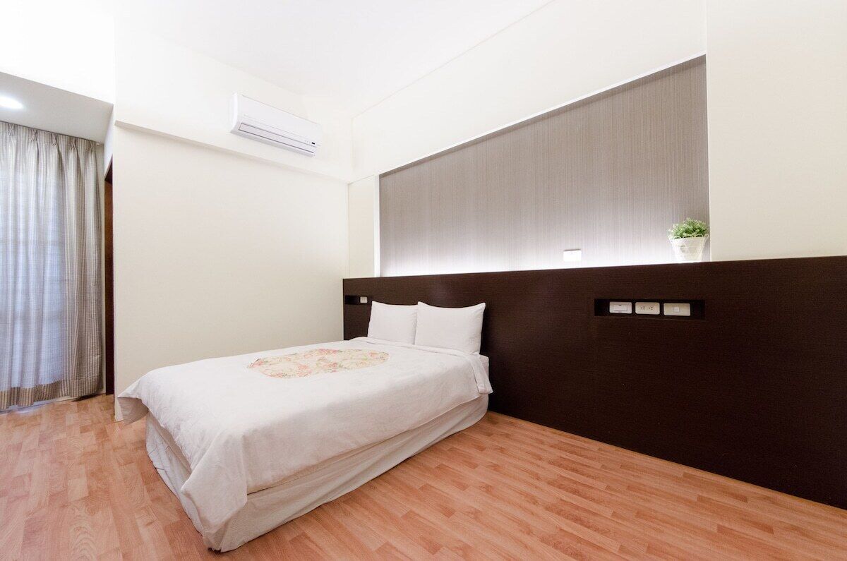 Habitación de hotel con cama, cabecera oscura, unidad de aire acondicionado, una planta pequeña y ventana con cortinas.