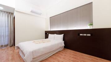 Habitación de hotel con cama, cabecera oscura, unidad de aire acondicionado, una planta pequeña y ventana con cortinas.