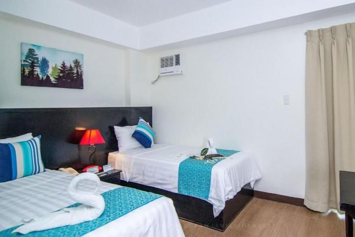 A hotel room with two beds, a desk, a chair, a painting, and an air conditioning unit.