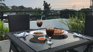 A table with two plates of food and two glasses of a dark beverage, set on a balcony with a view of the sky and distant buildings. - The Orient Beach Boracay (Boracay Island)