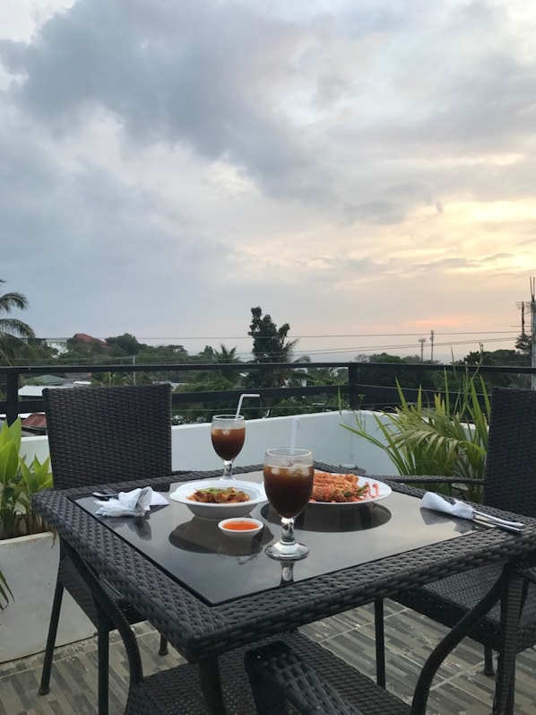 Une table avec deux assiettes de nourriture et deux verres d’une boisson sombre, placée sur un balcon d’où l’on peut admirer le ciel et des bâtiments éloignés.