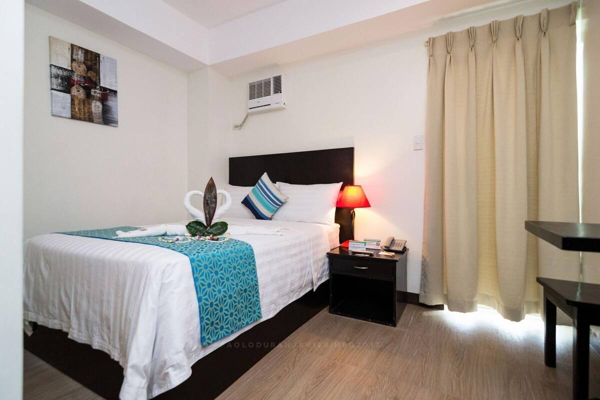 Een hotelkamer met een bed, nachtkastje, lamp, airconditioning en een schilderij aan de muur.