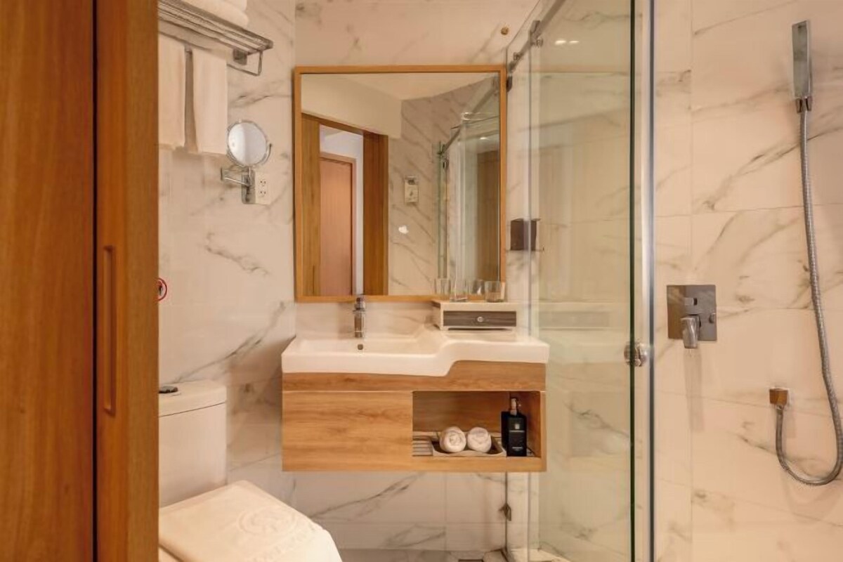 A modern bathroom with a marble wall, a wooden vanity, a glass shower enclosure, and a mirror above the sink.