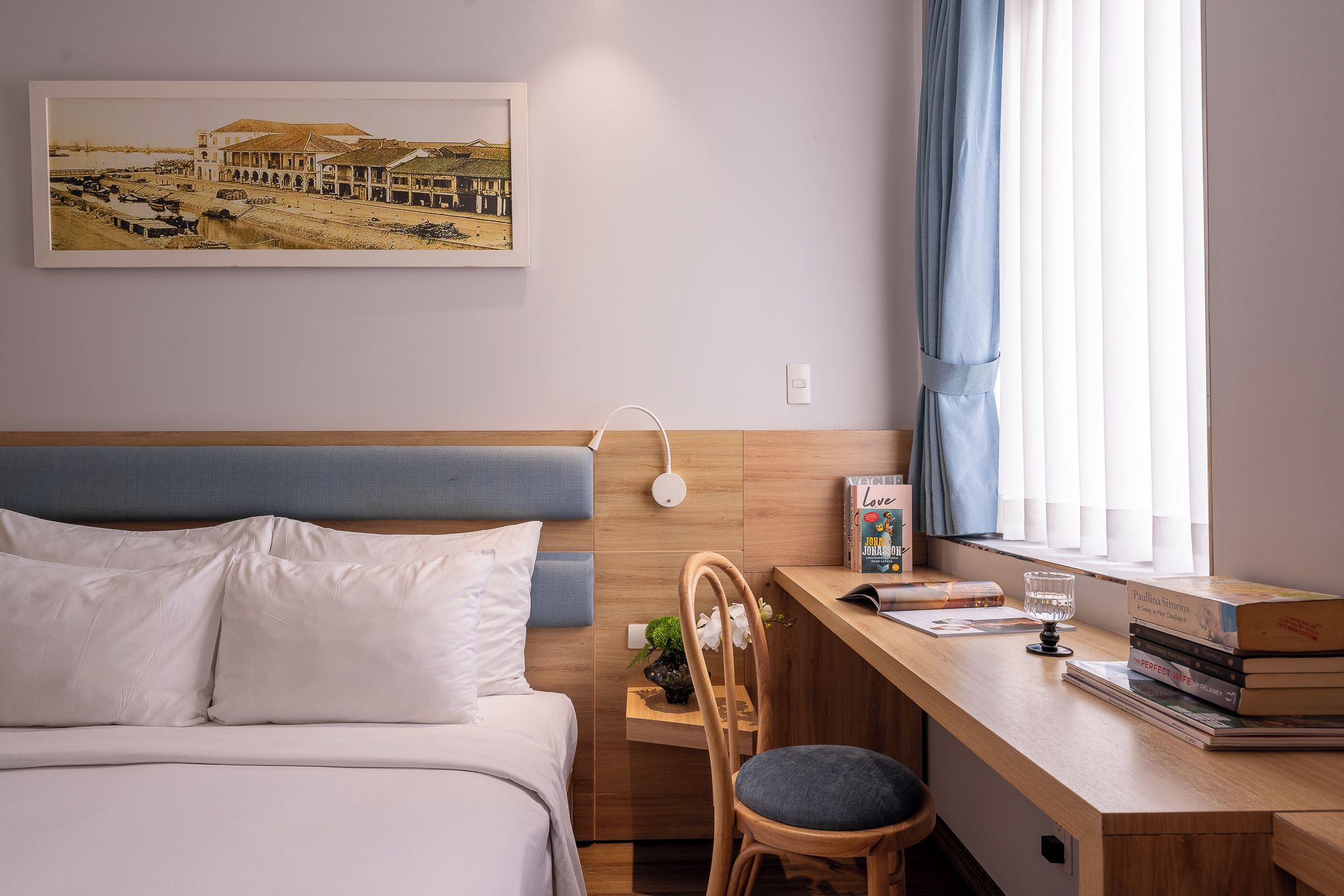 A hotel room with a bed, a desk, a chair, a window with curtains, a wall-mounted light, and a framed picture.